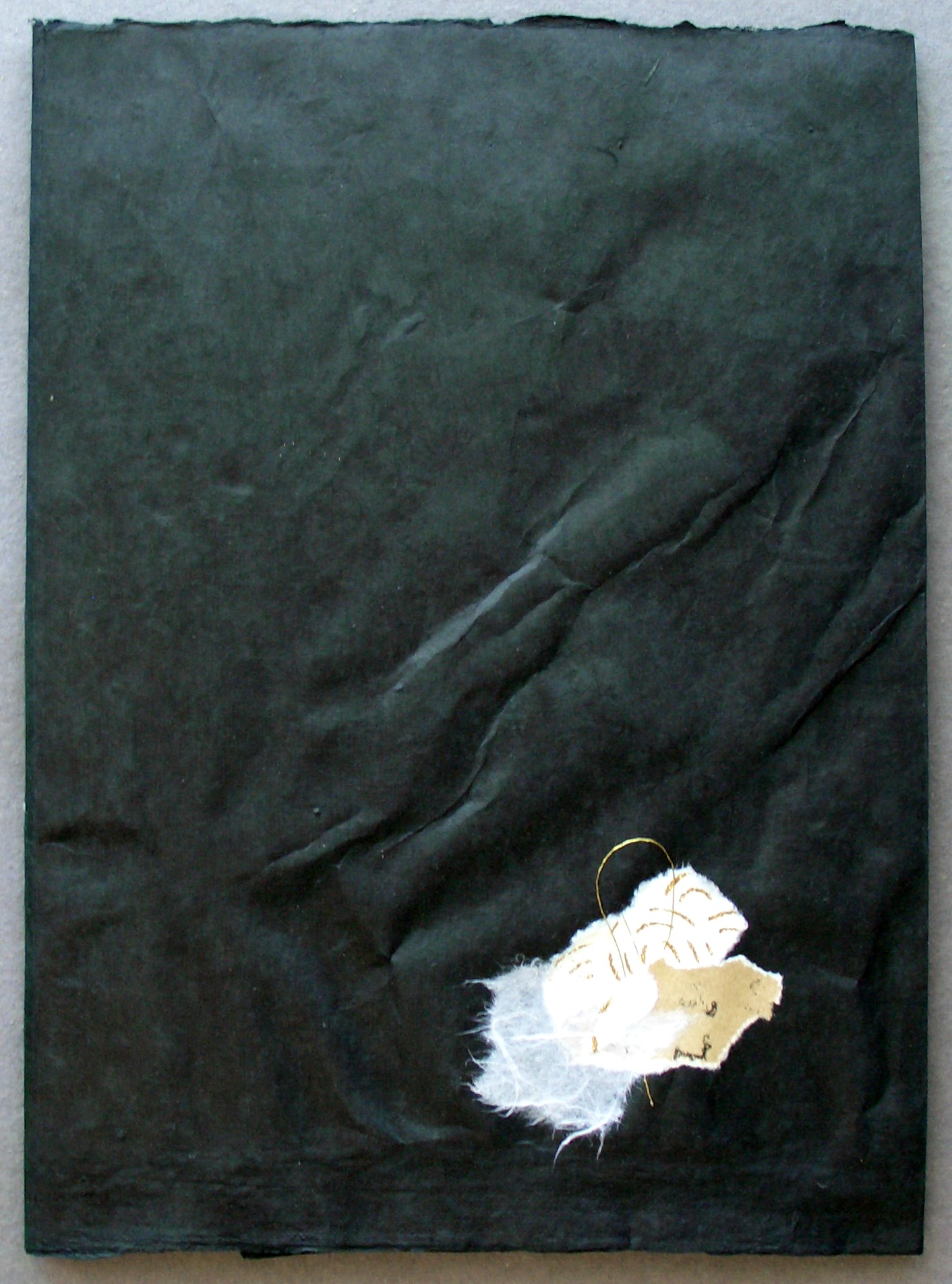 Black textured paper featuring a small, layered collage of white and beige fibers, accented with a gold paperclip. This artwork emphasizes innovative recycling in art.