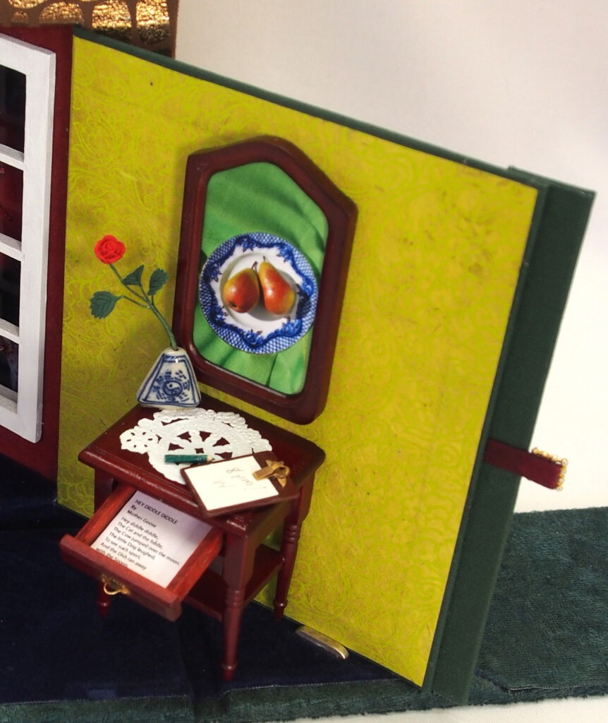 A detailed corner of a dollhouse showing a small table with a lace doily, a vase, and a fruit plate, set against a vibrant green wall.